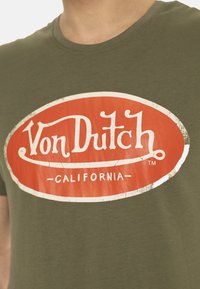 T-shirt olive à manches courtes avec un logo ovale rouge et du texte blanc "Von Dutch" et "CALIFORNIA", texture usée sur l'impression.
