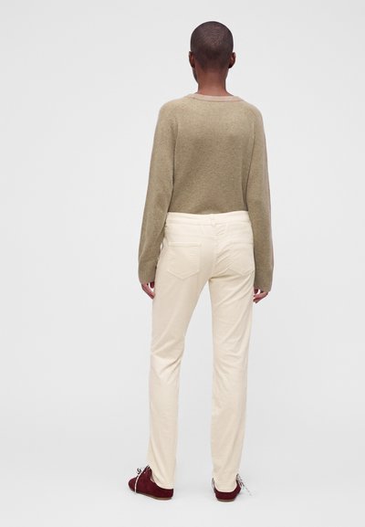 Personne aux cheveux courts portant un pull marron clair, un pantalon crème et des chaussures rouge foncé, debout de dos sur un fond uni.