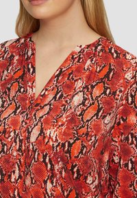 Rode en zwarte blouse met slangenprint, een diepe V-hals en een textuurstof, met oranje tinten in het patroon.