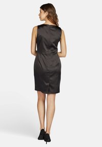 Schwarzes Satin-Kleid mit einer figurbetonten Silhouette, ärmellosem Design und einem Reißverschluss auf der Rückseite. Begleitet von schwarzen High-Heels.
