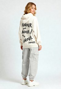 Felpa crema con testo nero "AMOUR", vestibilità rilassata, polsini a costine. Abbinata a pantaloni cargo grigi chiari, con caviglia elastica, e sneakers bianche.