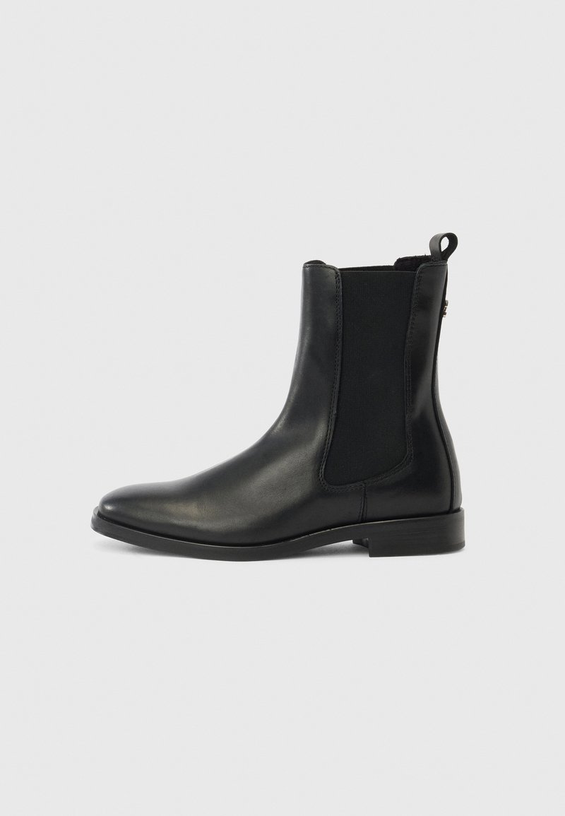 Bottine Chelsea en cuir noir avec panneau latéral élastique, talon bas, bout arrondi et languette arrière sur fond gris clair.
