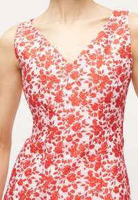 Αμάνικο φόρεμα με V λαιμόκοψη και κόκκινη κεντητική με λουλούδια σε λευκό ύφασμα, που δείχνει τη μέση και το επάνω μέρος του στήθους του φοράει.