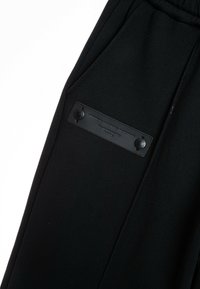 Pantalones jogger negros hechos de una tela suave, con un bolsillo lateral que cuenta con un parche de cuero suave y dos botones a presión negros.