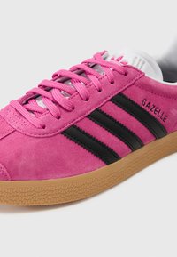 Pink suede sneaker with black leather stripes. Features flat pink laces, textured gum sole, and embossed "GAZELLE" branding on the side.