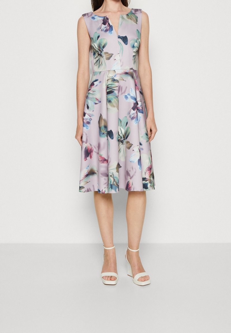Robe lavande sans manches longueur genou avec motifs floraux multicolores portée avec des sandales blanches à talons hauts.