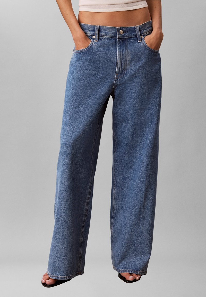 Μπλε τζιν ψηλόμεσο με φαρδιά μπατζάκια, φορεμένο με τα χέρια στις τσέπες και μαύρα σανδάλια ανοιχτής μύτης σε γκρι φόντο.