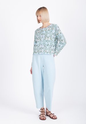 Vrouw die een patroonrijke langewerpen top draagt, lichtblauwe broek met brede pijpen en bruine sandalen met bandjes, staand tegen een witte achtergrond.