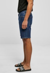 Shorts en denim bleu foncé avec une coupe droite, dotés d'une fermeture par bouton et zip, de poches latérales et d'une finition à cru. Portés avec des sandales noires à brides.