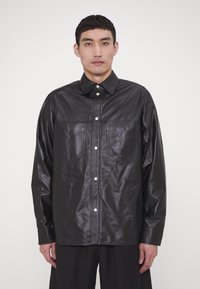 Chaqueta de camisa de cuero negro con cuello, cierre de botones a presión, dos bolsillos en el pecho y una textura suave.