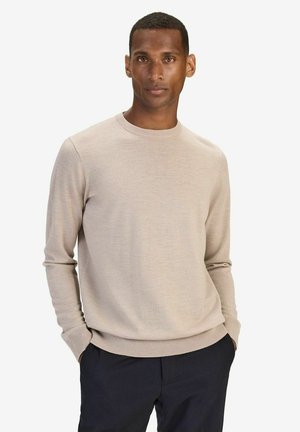 Beige gestrickter Pullover mit rundem Halsausschnitt und langen Ärmeln, der eine weiche Textur und eng anliegende Bündchen aufweist, kombiniert mit dunklen Hosen.