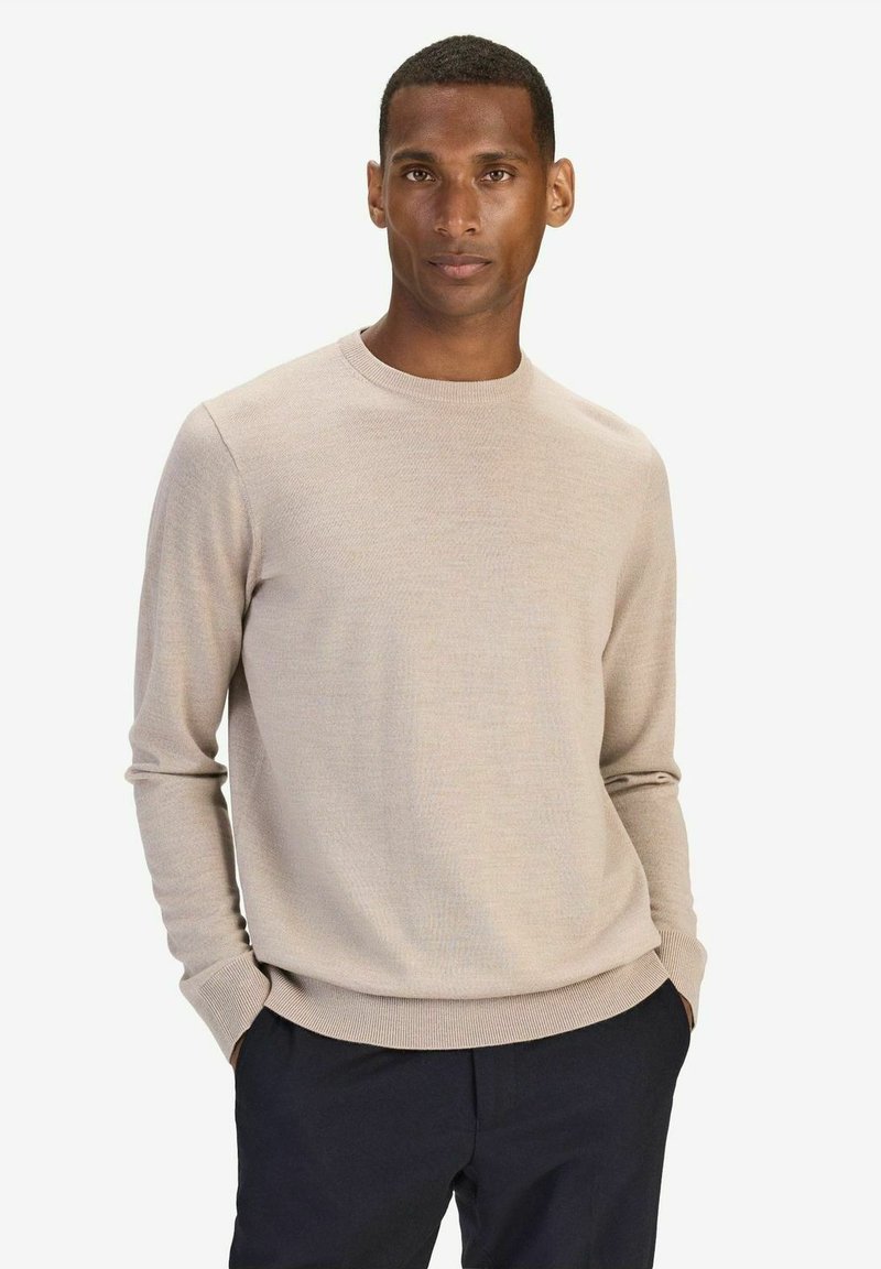 Beige stickad tröja med rund halsringning och långa ärmar, med en mjuk textur och åtsittande manschetter, parat med mörka byxor.