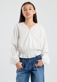 Blusa bianca tinta unita a maniche lunghe con scollo a V e orlo arricciato, abbinata a jeans in denim blu a vita alta.