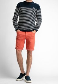 Pull rayé marine et blanc pour homme avec col rond, associé à un short corail retroussé. Des chaussures sombres sans lacets complètent le look.
