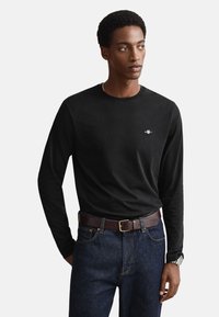 Camiseta negra de manga larga con escote redondo, material de algodón, detalle sutil de logo en el pecho, combinada con jeans de mezclilla oscuros y cinturón marrón.