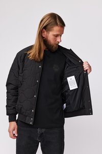 Veste matelassée noire avec des boutons-pression, poche intérieure et texture lisse. Portée sur une chemise noire avec un logo subtil.