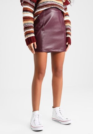 Kvinde iført en burgundy lædernederdel, stribet striktrøje og hvide høje sneakers, stående mod en ensfarvet baggrund.