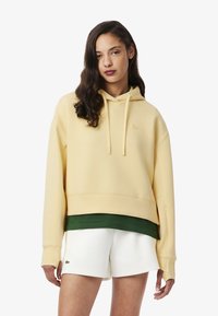 Gelbe Kapuzenjacke mit lockerer Passform, ausgestattet mit einer Kängurutasche, geripptem Saum und einem kleinen Logo. Kombiniert mit weißen Shorts.