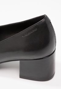 Chaussure à talon haut en cuir noir avec un talon carré, texture lisse et logo "VAGABOND" en relief sur le côté. Design minimaliste.