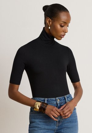 Femme avec les cheveux en chignon portant un col roulé noir à manches courtes, un jean bleu, des bracelets en or et en écaille de tortue, et de petites boucles d'oreilles rondes regardant vers le bas.