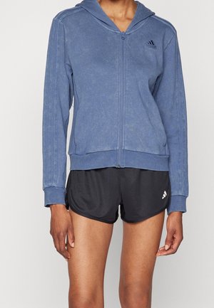 Personne portant un sweat à capuche zippé bleu avec logo et un short de sport noir avec logo, debout devant un fond uni.