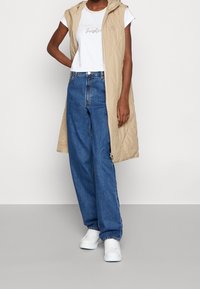 Beige quilted vest med hætte, lynlåslukning og logo detalje. Parret med en hvid T-shirt og blå højtaljede jeans. Hvide sneakers.