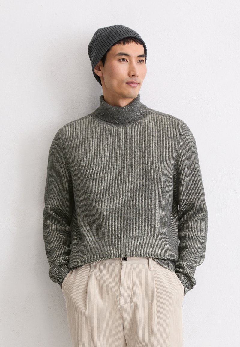 Uomo che indossa un maglione a collo alto a coste grigio, pantaloni beige e un berretto grigio scuro, con le mani in tasca appoggiato a un muro bianco.
