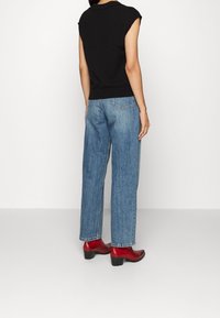 Schwarzes ärmelloses Top, blaue gerade geschnittene Jeans und rote Absatzstiefel bis zum Knöchel, von hinten vor einem schlichten weißen Hintergrund betrachtet.