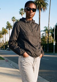 Persoon met zwarte zonnebril, zwarte capuchonjas en lichtgekleurde spijkerbroek, staand met handen in de zakken op een zonnige straat omzoomd met palmbomen.