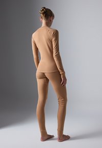 Woman stands barefoot with back turned, wearing fitted tan long-sleeve top and matching leggings against neutral background.