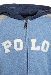 Světle modrá mikina s kapucí na zip, šedé a námořnické panely a bílé plstěné písmena s nápisem "POLO" na přední straně. Měkká textilie.