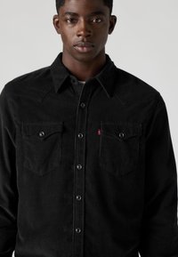 Camisa preta de veludo cotelê com botões de pressão, dois bolsos no peito e gola apontada. Detalhes notáveis de costura nos ombros.