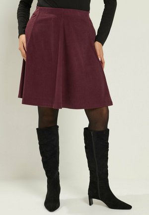 Mujer que lleva una falda burdeos de pana con corte en A, medias negras, botas negras altas hasta la rodilla con tacón, y una camiseta negra de manga larga, de pie con las manos en los bolsillos.