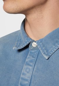 Camicia in corduroy blu con colletto a punta, chiusura con bottoni e sottili righe verticali in una texture morbida, che mette in evidenza cuciture dettagliate.