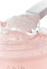 Prodotto in gel in un contenitore trasparente, caratterizzato da una tonalità rosa chiaro con bolle e una consistenza liscia. Un applicatore bianco è parzialmente visibile.