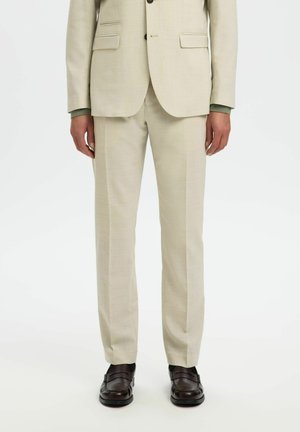 Helle beigefarbene Anzug mit texturiertem Finish, ausgestattet mit einem einreihigen Jacket und maßgeschneiderten Hosen, ergänzt durch dunkelbraune Schuhe.
