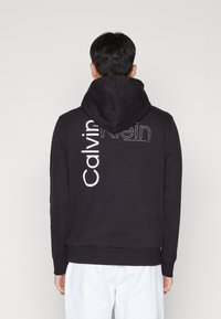 Černá mikina s kapucí vyrobená z bavlny, na zadní straně s velkým bílým potiskem "Calvin Klein". Obsahuje velkou nastavitelnou kapuci a žebrované manžety.