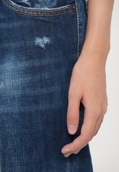 Jeans de mezclilla azul oscuro con un sutil detalle desgastado en forma de corazón. La tela presenta un acabado texturizado, con costuras de color naranja en contraste.