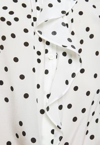 Gros plan sur un tissu blanc à pois noirs, avec un bord froncé et une fermeture à boutons blancs en dessous.