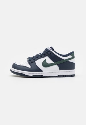 Baskets basses avec base blanche, surpiqûres bleu foncé, logo Nike vert foncé et lacets bleu foncé sur fond blanc.