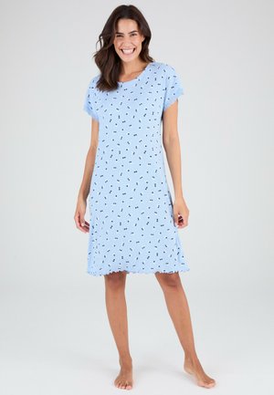 Lächelnde Frau, barfuß, in einem hellblauen, kurzärmeligen, knielangen Kleid mit kleinen dunklen Mustern vor schlichtem Hintergrund.