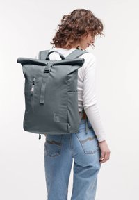 Sac à dos gris à rabat avec une poche zippée à l'avant, des bretelles réglables et un logo de marque discret sur le côté. Fabriqué en matériau durable.