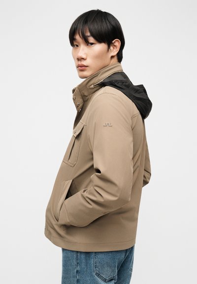 Jeune homme portant une veste beige avec une capuche noire et un jean bleu, les mains dans les poches, regardant par-dessus son épaule sur un fond uni.