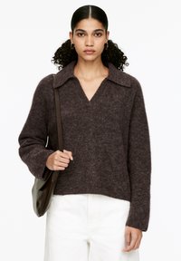 Frau mit lockigem Haar, die Ohrringe in Form von Ringen trägt, einen dunkelbraunen, flauschigen Pullover, weiße Hosen und eine braune Schultertasche trägt und vor einem weißen Hintergrund steht.