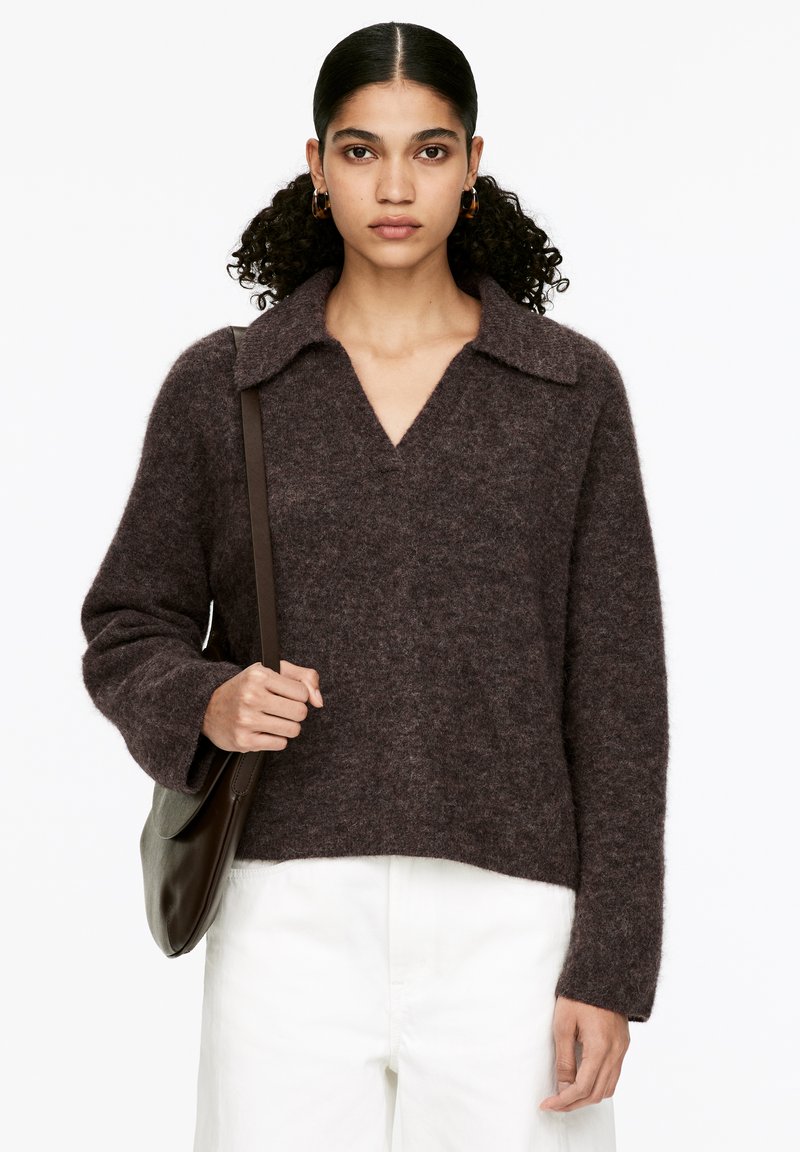 Frau mit lockigem Haar, die Ohrringe in Form von Ringen trägt, einen dunkelbraunen, flauschigen Pullover, weiße Hosen und eine braune Schultertasche trägt und vor einem weißen Hintergrund steht.