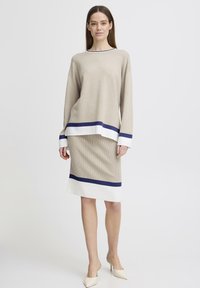 Femme portant un pull en tricot beige et une jupe à côtes assortie avec des rayures blanches et bleues, se tenant sur des talons beiges sur un fond blanc.