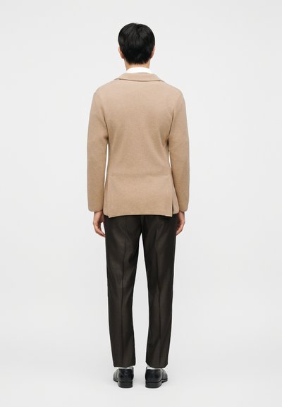 Μπεζ πλεκτή ζακέτα με γιακά σάλι, λεπτομέρεια πίσω άνοιγμα, σε συνδυασμό με σκούρο, υφασμάτινο παντελόνι και μαύρα παπούτσια.