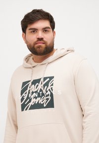Hoodie w jasnym beżu, wykonany z miękkiego materiału. Posiada czarno-biały graficzny logo z przodu oraz regulowane sznurki.