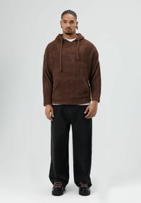 Sweat à capuche marron et duveteux avec une poche avant et une capuche à cordon, associé à un jean noir ample et des chaussures noires à lacets roses.