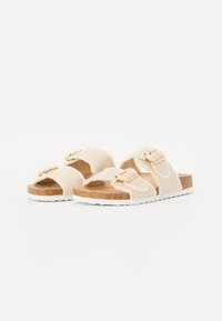 Beige, fuzzy slidesandaler med to justerbare stropper, gullfargede detaljer, korke midtsåle og hvit teksturert gummisåle.